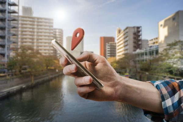 Person using smartphone with location pin icon in a modern city near waterfront buildings.